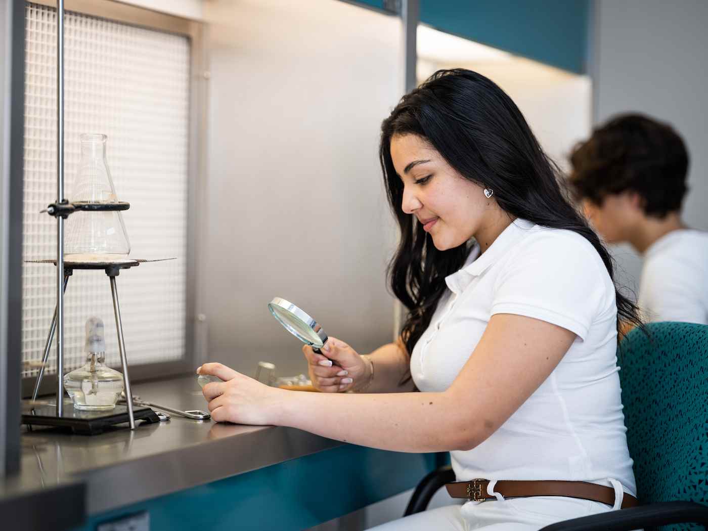 Estudiante observa una muestra con lupa en un laboratorio, junto a un matraz sobre un mechero Bunsen.